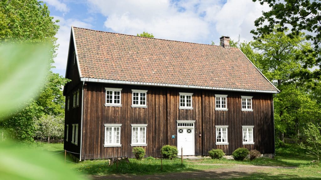 Gammelt trehus med mørk fasade og rødt takstein, omgitt av grønt gress og trær.