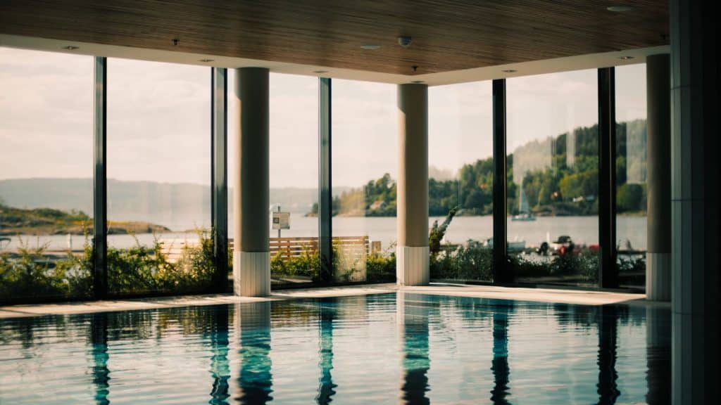 Innendørs basseng med store vinduer som gir utsikt mot innsjø, trær og åser i bakgrunnen