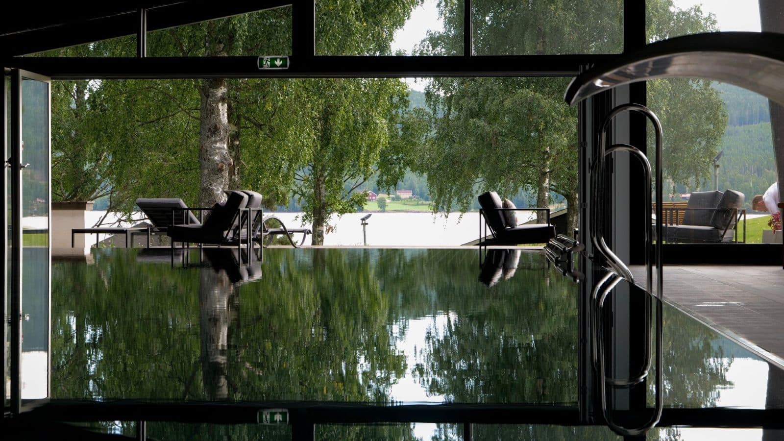 Innendørs basseng med store glassvinduer som speiler trær og utemøbler, omgitt av lounge-stoler og utsikt mot naturen