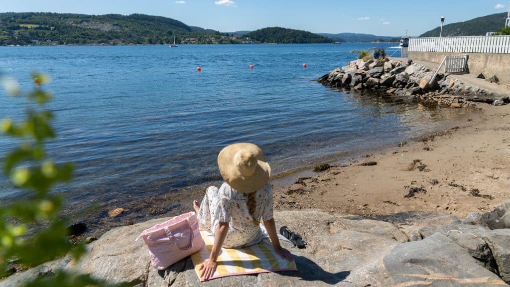 Dame med hatt som sitter på et strandhåndkle og ser utover vannet