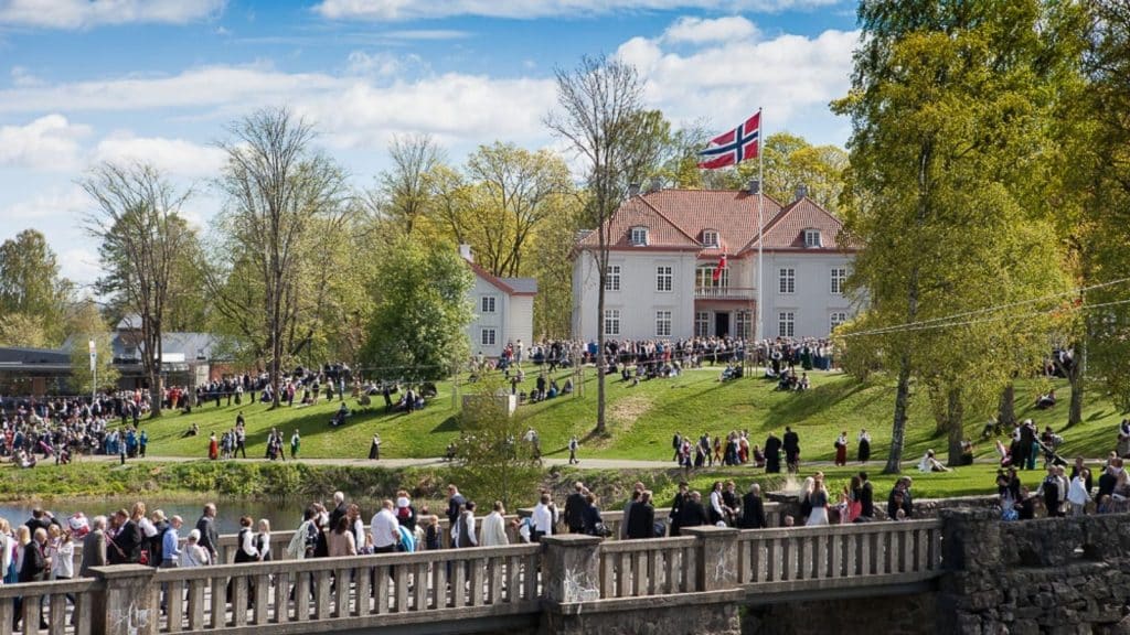 Folk samles foran Eidsvollsbygningen 17.mai med norske flagg, omgitt av grønt landskap og historisk arkitektur