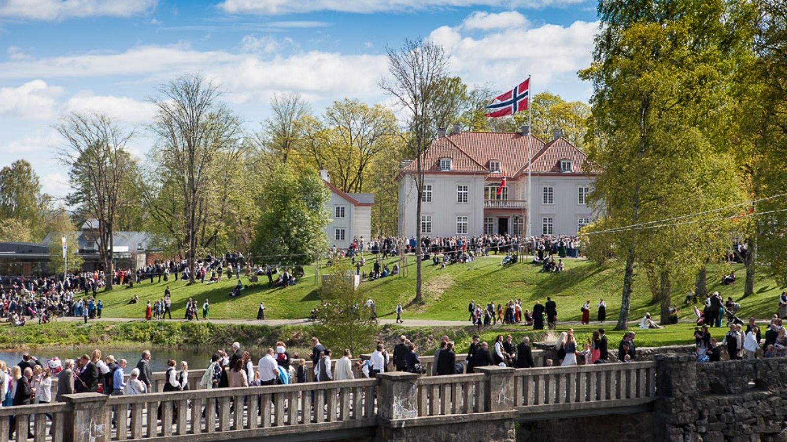 Folk samles foran Eidsvollsbygningen 17.mai med norske flagg, omgitt av grønt landskap og historisk arkitektur