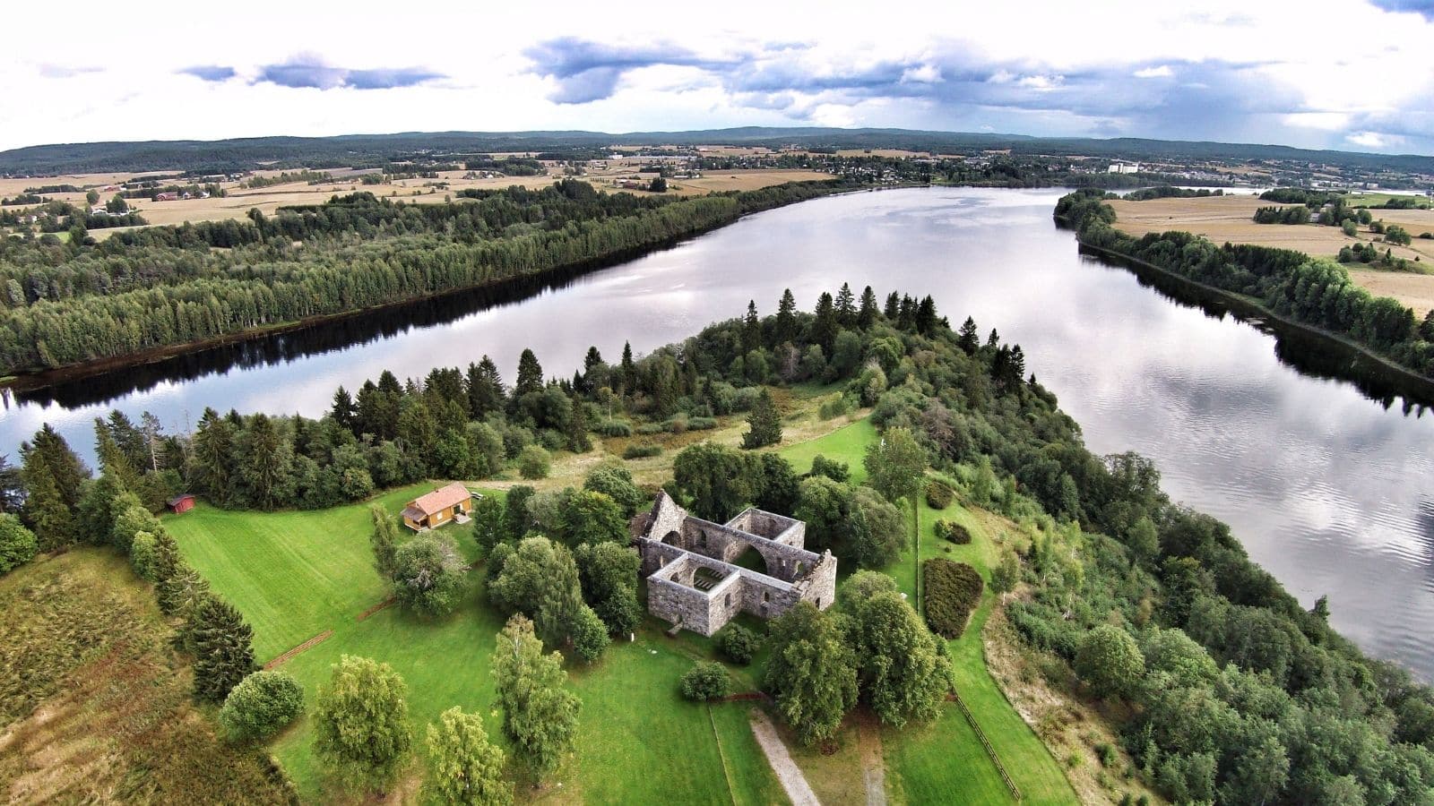 Sommerbilde tatt med Drone av Nes kirkeruiner omgitt av grønn natur og elvemøtet Glomma og Vorma på Romerike