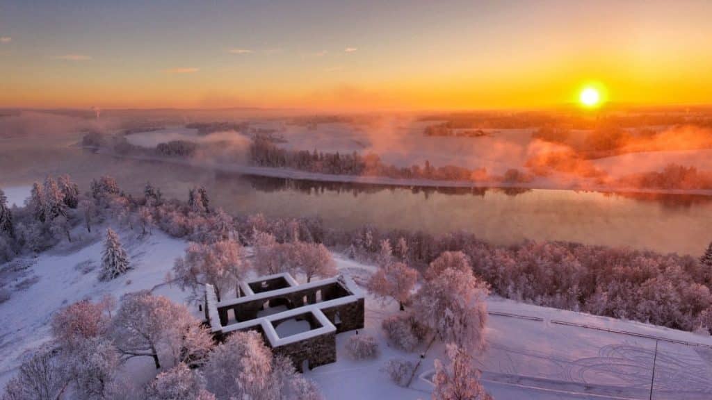 Nes kirkeruiner i vinterlandskap med snø, solnedgang og elvemøtet i Nes på Romerike