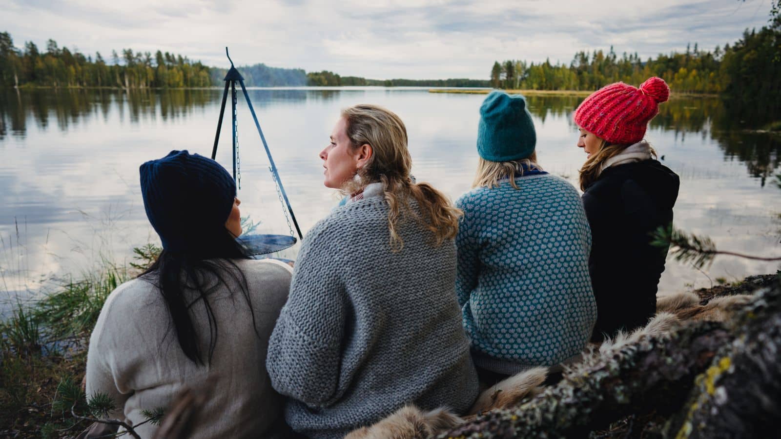 To personer sitter på en stein ved vannkanten med utsikt over en innsjø og skog i bakgrunnen. En kano med norske flagg ligger ved bryggen
