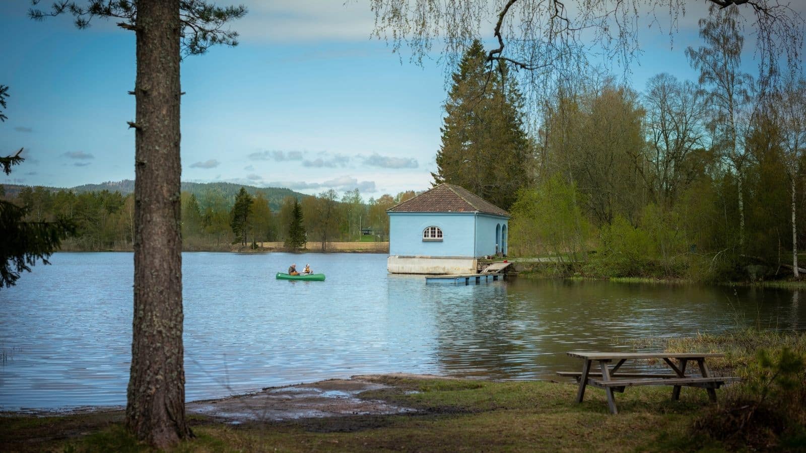 Blått hus ved en liten sjø med to personer i en kajakk på vannet og skog rundt